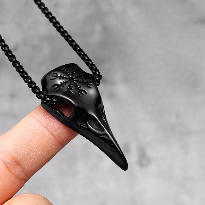 Pendentif crâne de corbeau noir suspendu à une chaîne tenu entre les doigts sur fond gris